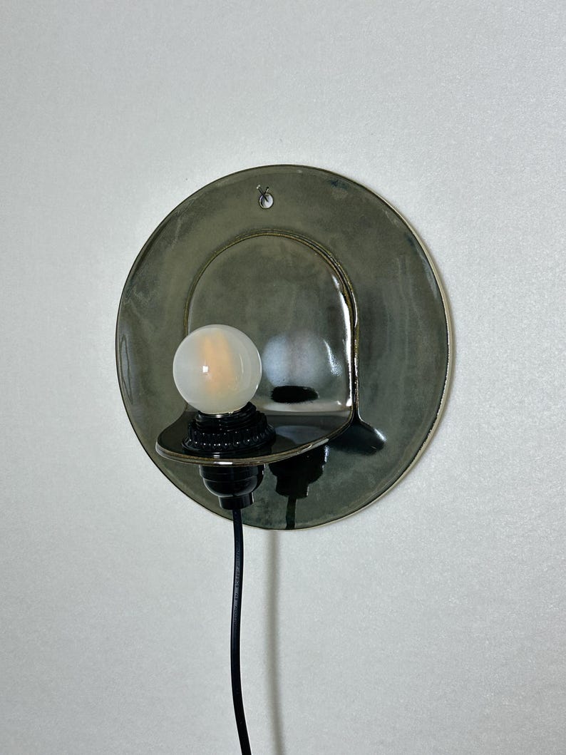 May include: A wall-mounted sconce featuring a circular, glossy, dark olive-green ceramic backplate with a small hole at the top. A black socket holds a round, frosted white light bulb, and a thin black power cord extends downward from the base.