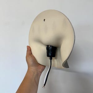 May include: A cream-colored, semi-circular wall lamp fixture with a smooth, curved surface. The center features a black socket for a light bulb, with a black and white electrical cord extending downward. A small mounting hole is visible at the top.