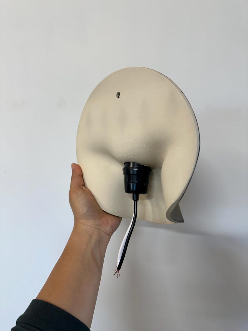 May include: A cream-colored, semi-circular wall lamp fixture with a smooth, curved surface. The center features a black socket for a light bulb, with a black and white electrical cord extending downward. A small mounting hole is visible at the top.