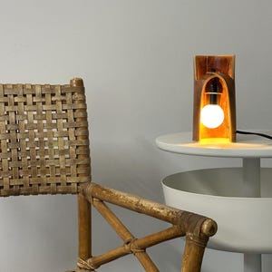 Puede incluir: Una l&aacute;mpara de mesa cil&iacute;ndrica con un exterior marr&oacute;n c&aacute;lido y un interior brillante, que presenta una bombilla blanca expuesta. La l&aacute;mpara est&aacute; colocada sobre una superficie blanca junto a una silla de rat&aacute;n tejida con marco cruzado.