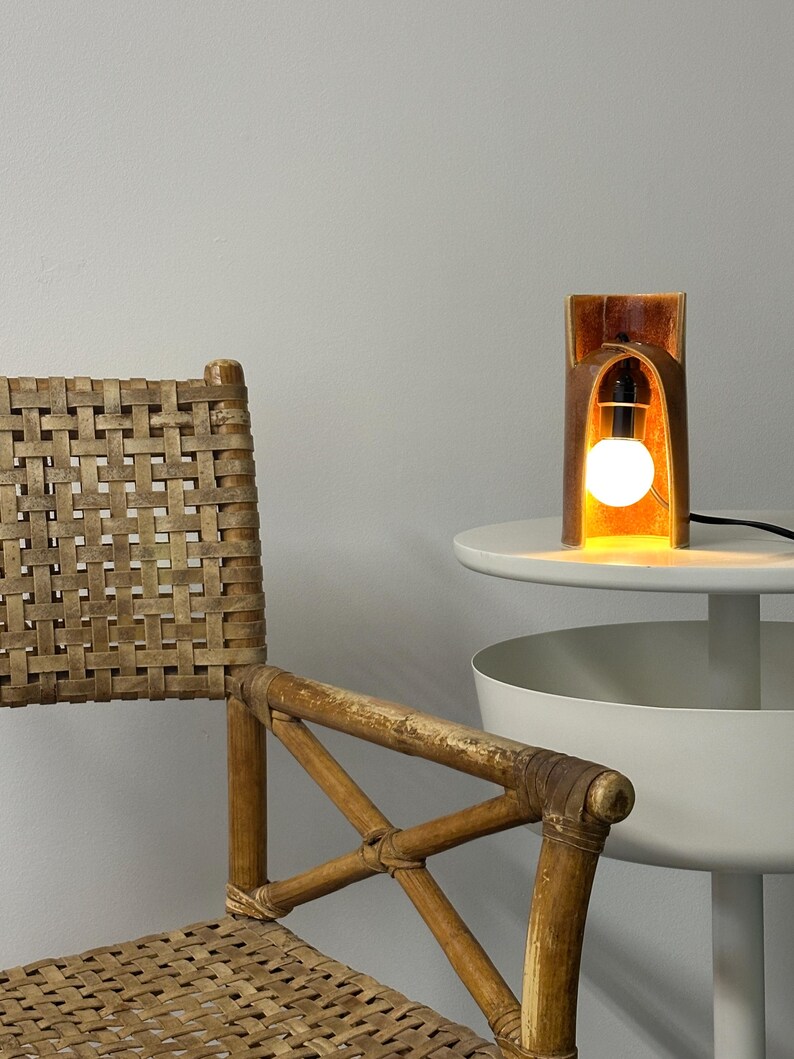 Puede incluir: Una l&aacute;mpara de mesa cil&iacute;ndrica con un exterior marr&oacute;n c&aacute;lido y un interior brillante, que presenta una bombilla blanca expuesta. La l&aacute;mpara est&aacute; colocada sobre una superficie blanca junto a una silla de rat&aacute;n tejida con marco cruzado.