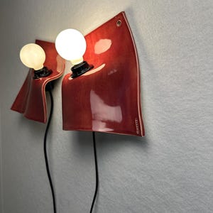 Puede incluir: Dos l&aacute;mparas de pared con pantallas de cer&aacute;mica de color rojo intenso, curvas y brillantes, con un acabado liso. Cada l&aacute;mpara tiene un casquillo negro con una bombilla blanca redonda y brillante, y un cable negro hacia abajo.