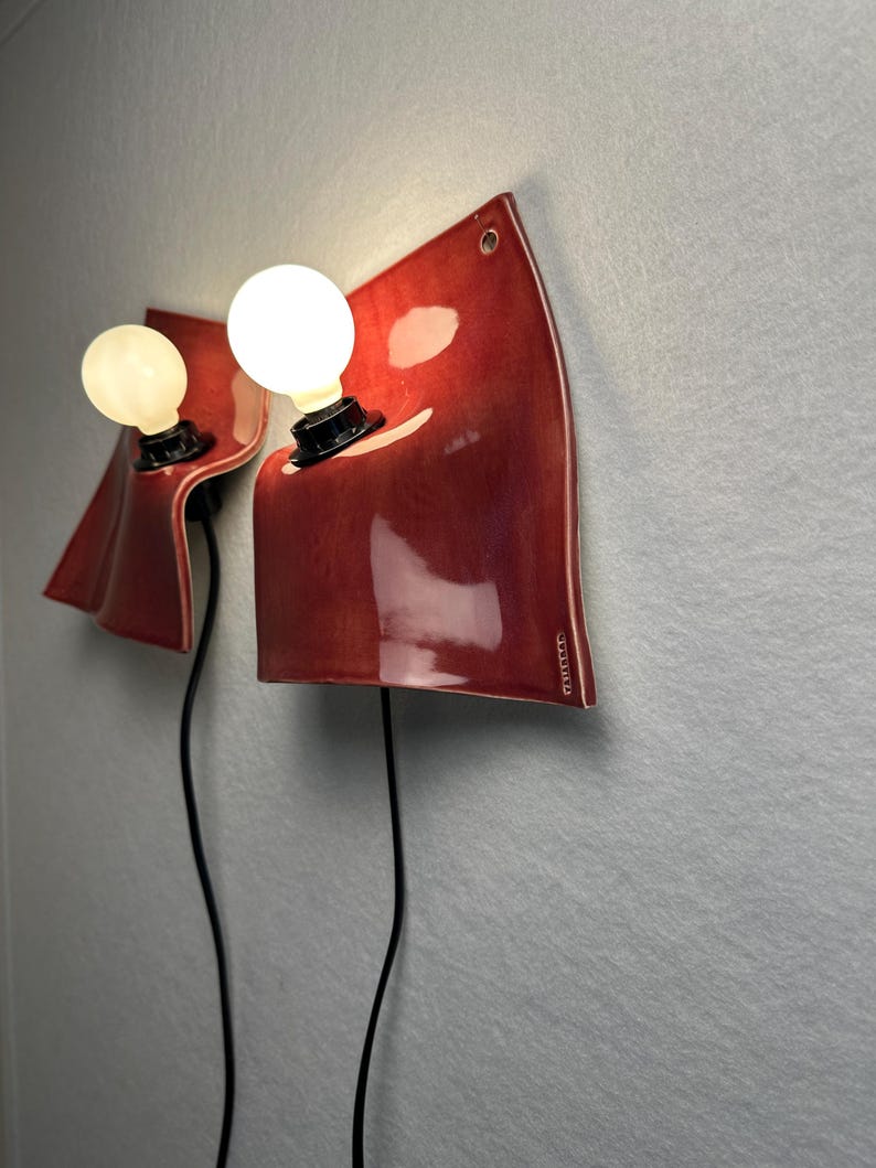 Puede incluir: Dos l&aacute;mparas de pared con pantallas de cer&aacute;mica de color rojo intenso, curvas y brillantes, con un acabado liso. Cada l&aacute;mpara tiene un casquillo negro con una bombilla blanca redonda y brillante, y un cable negro hacia abajo.