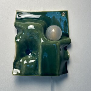 May include: A wall-mounted ceramic light fixture with a glossy, undulating olive green surface. The piece features two circular holes at the top for mounting and a single, spherical white glass bulb integrated into the molded, wavy form.