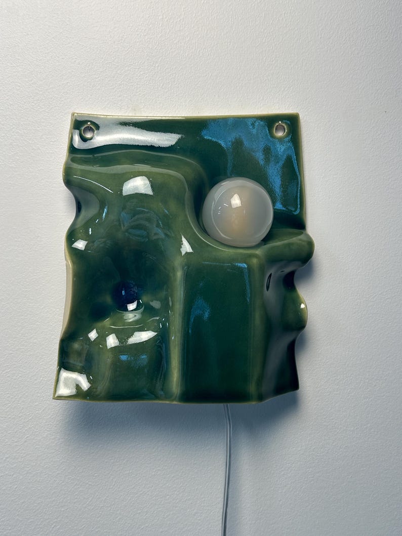 May include: A wall-mounted ceramic light fixture with a glossy, undulating olive green surface. The piece features two circular holes at the top for mounting and a single, spherical white glass bulb integrated into the molded, wavy form.