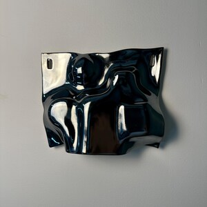 Pu&ograve; includere: Una scultura da parete in ceramica blu scuro lucida con una superficie irregolare e ondulata. Il pezzo presenta una forma fluida simile a un'onda con curve profonde e riflessi. Include due fori di montaggio rettangolari in alto.