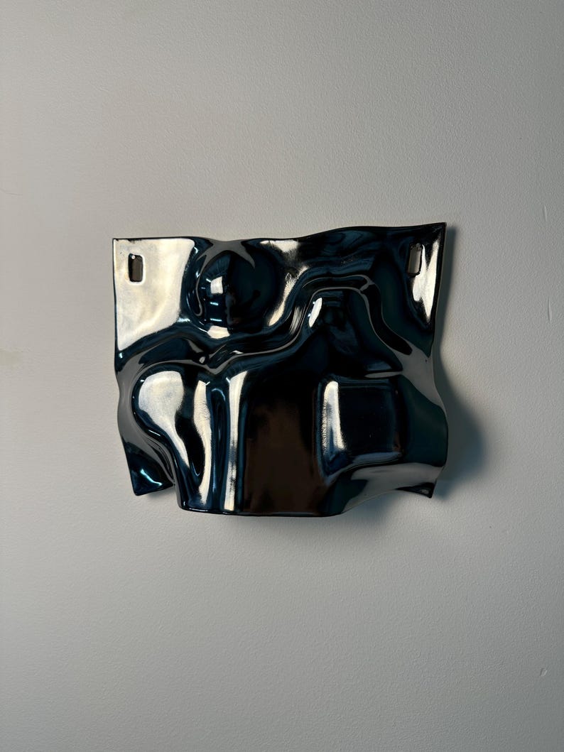 Pu&ograve; includere: Una scultura da parete in ceramica blu scuro lucida con una superficie irregolare e ondulata. Il pezzo presenta una forma fluida simile a un'onda con curve profonde e riflessi. Include due fori di montaggio rettangolari in alto.