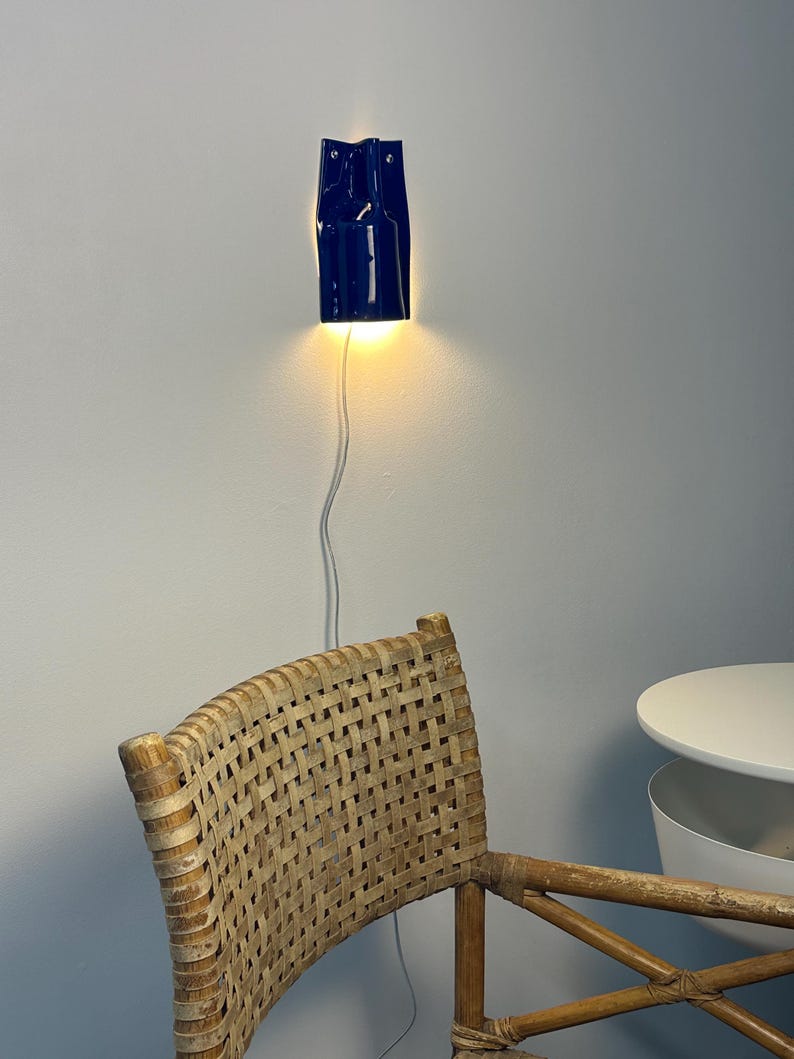 Puede incluir: Un aplique de pared de cer&aacute;mica azul oscuro brillante con un dise&ntilde;o vertical y plegado. La l&aacute;mpara est&aacute; montada en una pared y emite una luz c&aacute;lida dirigida hacia abajo. Un cable de alimentaci&oacute;n blanco y fino cuelga desde la base.