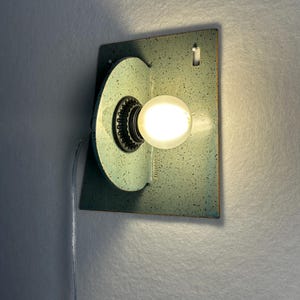 May include: A wall-mounted light fixture featuring a rectangular backplate and a perpendicular circular element, both finished in a speckled light blue-green glaze. A single glowing round light bulb is centered in the circular component.