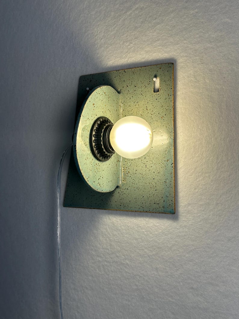 May include: A wall-mounted light fixture featuring a rectangular backplate and a perpendicular circular element, both finished in a speckled light blue-green glaze. A single glowing round light bulb is centered in the circular component.