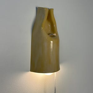 Pu&ograve; includere: Un'applique da parete in ceramica con una finitura lucida color giallo pallido. L'apparecchio presenta una forma organica alta e curva con un'apertura centrale e una lampadina che emette una luce calda dal basso, collegata da un cavo trasparente.