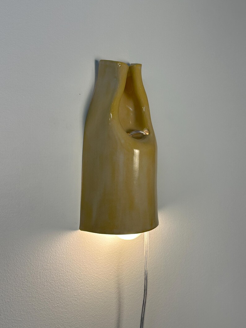 Pu&ograve; includere: Un'applique da parete in ceramica con una finitura lucida color giallo pallido. L'apparecchio presenta una forma organica alta e curva con un'apertura centrale e una lampadina che emette una luce calda dal basso, collegata da un cavo trasparente.