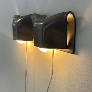 Könnte beinhalten: Eine Wandleuchte mit zwei Köpfen, bestehend aus zwei dunklen, geschwungenen Metallschirmen mit glatter Oberfläche. Jeder Schirm enthält eine warm leuchtende Glühbirne, die das Licht nach unten wirft. Dünne, transparente Stromkabel hängen von den Leuchten.