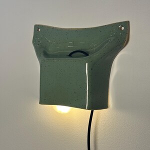 May include: A wall-mounted ceramic light fixture in a glossy sage green finish with dark speckles. The piece features a curved, flared top with two mounting holes and a rectangular base that emits a warm glow from a light bulb underneath.