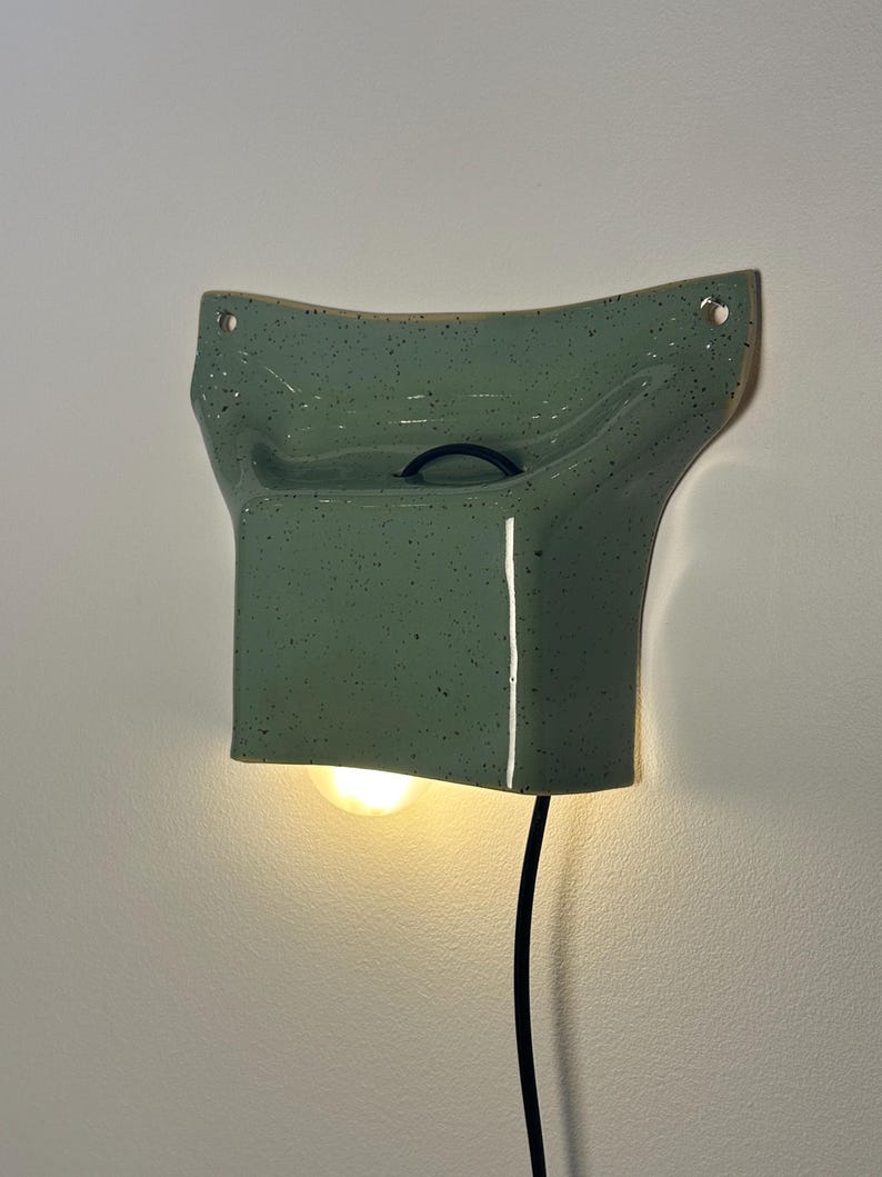 May include: A wall-mounted ceramic light fixture in a glossy sage green finish with dark speckles. The piece features a curved, flared top with two mounting holes and a rectangular base that emits a warm glow from a light bulb underneath.