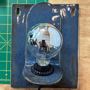 May include: A wall-mounted ceramic light fixture featuring a glossy, deep blue rectangular backplate with two rectangular cutouts at the top. A black circular base holds a large, clear, spherical glass light bulb with visible internal filaments.