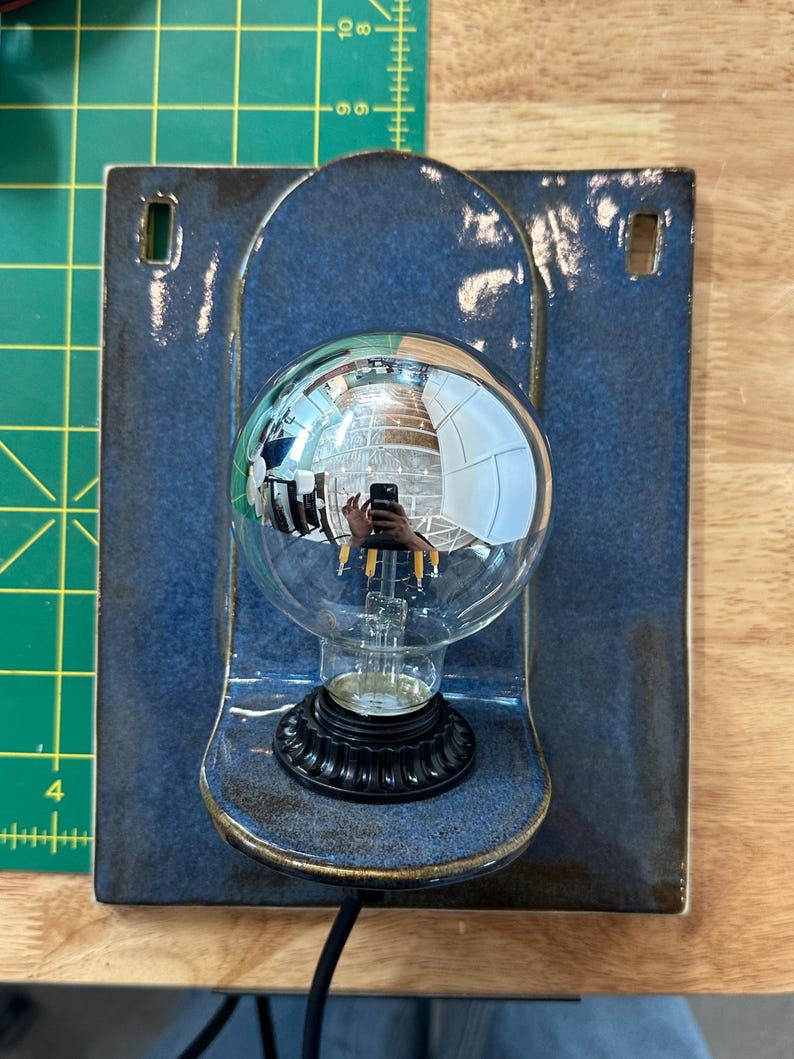 May include: A wall-mounted ceramic light fixture featuring a glossy, deep blue rectangular backplate with two rectangular cutouts at the top. A black circular base holds a large, clear, spherical glass light bulb with visible internal filaments.