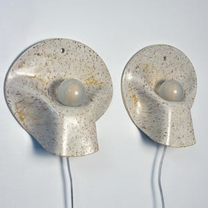 May include: A pair of wall-mounted ceramic sconces featuring a speckled cream and brown finish. Each lamp has a circular, undulating form with a central, spherical frosted glass bulb and a clear electrical cord extending from the bottom.