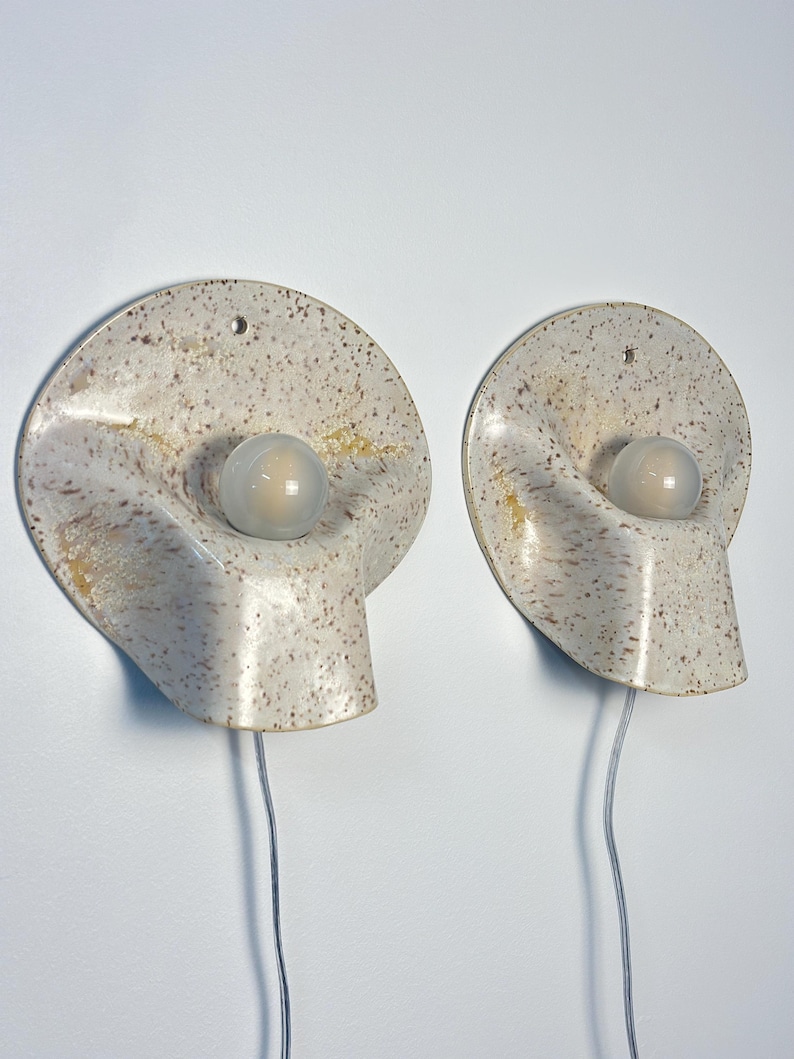 May include: A pair of wall-mounted ceramic sconces featuring a speckled cream and brown finish. Each lamp has a circular, undulating form with a central, spherical frosted glass bulb and a clear electrical cord extending from the bottom.