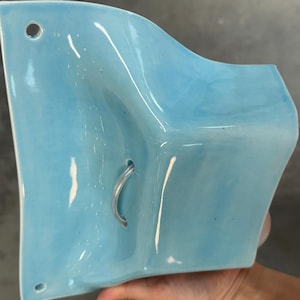 K&ouml;nnte beinhalten: Eine hellblaue Wandtasche aus Keramik mit gl&auml;nzender Oberfl&auml;che und geschwungener, skulpturaler Form. Das St&uuml;ck verf&uuml;gt &uuml;ber zwei kleine kreisf&ouml;rmige L&ouml;cher oben und unten zur Befestigung sowie eine mittlere &Ouml;ffnung mit einem kleinen, gebogenen Metallhaken.