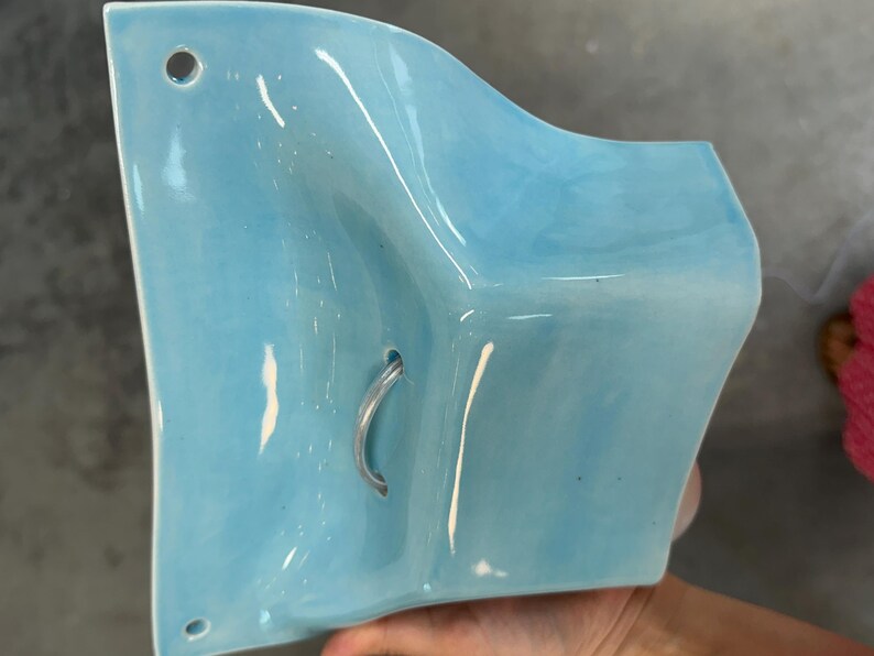 K&ouml;nnte beinhalten: Eine hellblaue Wandtasche aus Keramik mit gl&auml;nzender Oberfl&auml;che und geschwungener, skulpturaler Form. Das St&uuml;ck verf&uuml;gt &uuml;ber zwei kleine kreisf&ouml;rmige L&ouml;cher oben und unten zur Befestigung sowie eine mittlere &Ouml;ffnung mit einem kleinen, gebogenen Metallhaken.
