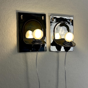 May include: Two wall-mounted sconces with a dark, glossy finish. Each sconce features a curved, reflective interior and a round, illuminated light bulb. The sconces are connected to a wall outlet via a thin, silver cord, offering a modern lighting solution.
