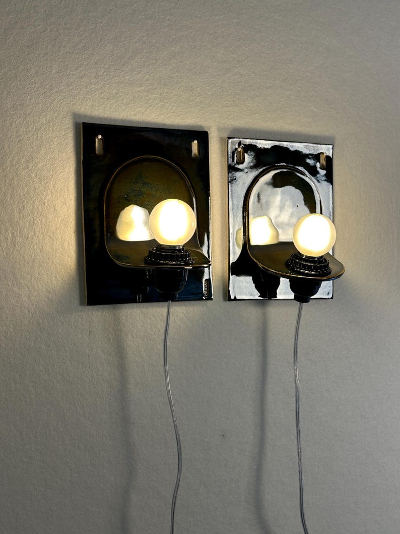 May include: Two wall-mounted sconces with a dark, glossy finish. Each sconce features a curved, reflective interior and a round, illuminated light bulb. The sconces are connected to a wall outlet via a thin, silver cord, offering a modern lighting solution.