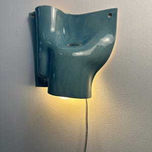 May include: A wall-mounted light fitting with a glossy, teal-coloured ceramic shade featuring an organic, undulating form. The fitting includes two circular mounting holes at the top corners and emits a warm, downward-facing glow from a bulb.