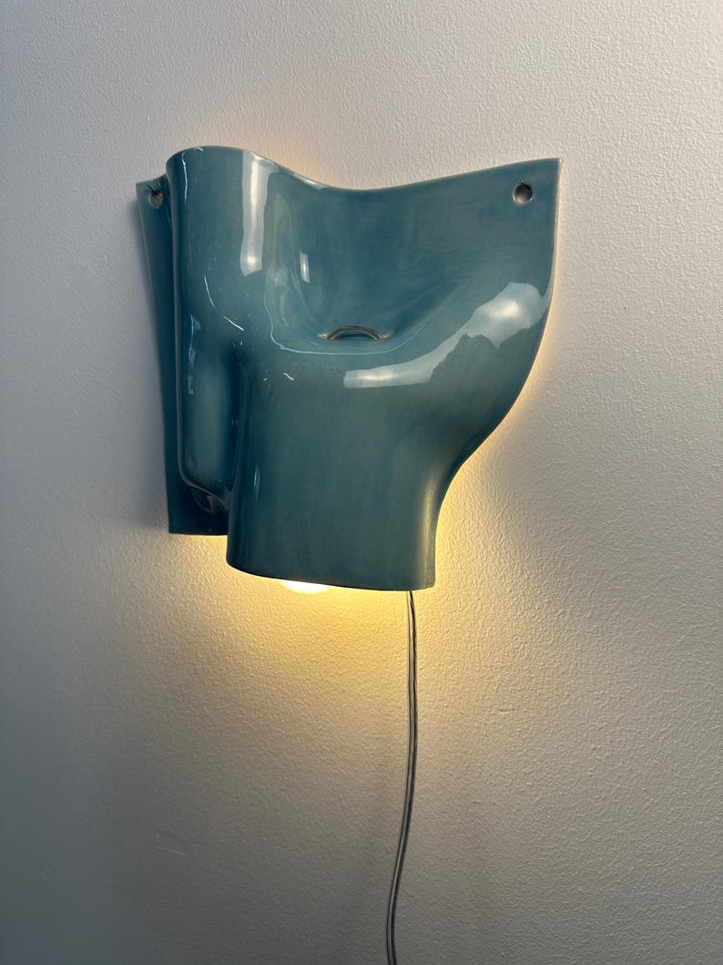 May include: A wall-mounted light fitting with a glossy, teal-coloured ceramic shade featuring an organic, undulating form. The fitting includes two circular mounting holes at the top corners and emits a warm, downward-facing glow from a bulb.