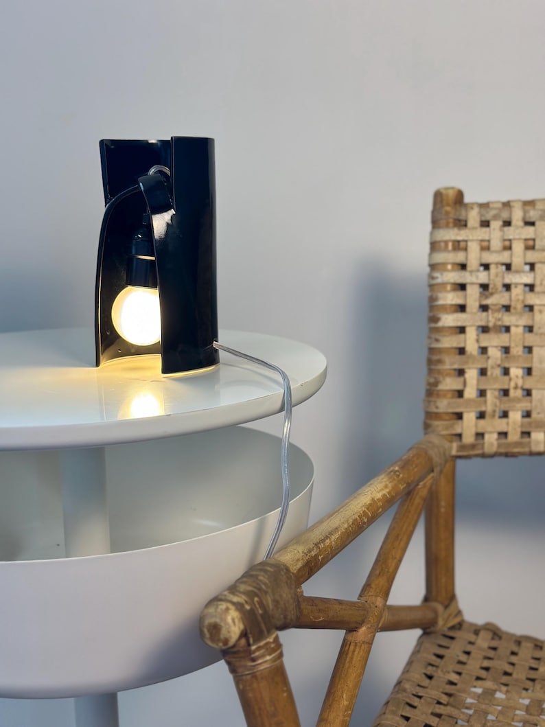 May include: A black cylindrical table lamp with a glossy finish and an open front section. The lamp features a single exposed light bulb emitting a warm glow, connected to a thin, clear power cord that extends from the base of the fixture.