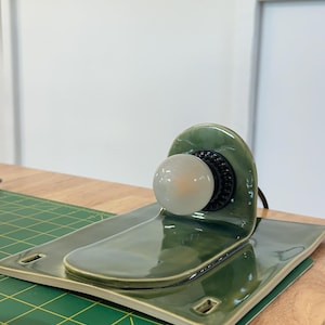 May include: A glossy olive green ceramic lamp base with a curved back panel and a flat rectangular platform. A black socket is mounted on the vertical section, holding a small, round, frosted white light bulb. The ceramic has a smooth finish.