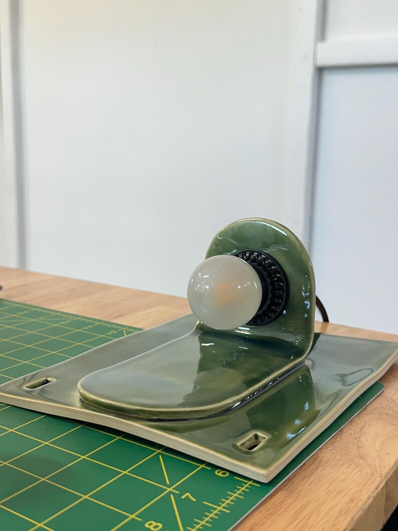 May include: A glossy olive green ceramic lamp base with a curved back panel and a flat rectangular platform. A black socket is mounted on the vertical section, holding a small, round, frosted white light bulb. The ceramic has a smooth finish.
