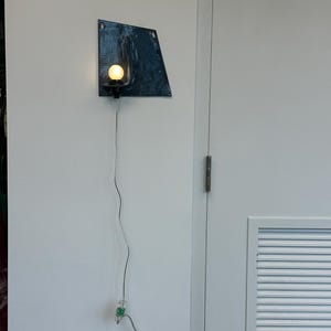 May include: A modern wall sconce with a dark blue, irregularly shaped metal backplate. A small, round light bulb is mounted on a black fixture. The sconce is connected to a wall outlet via a gray cord.