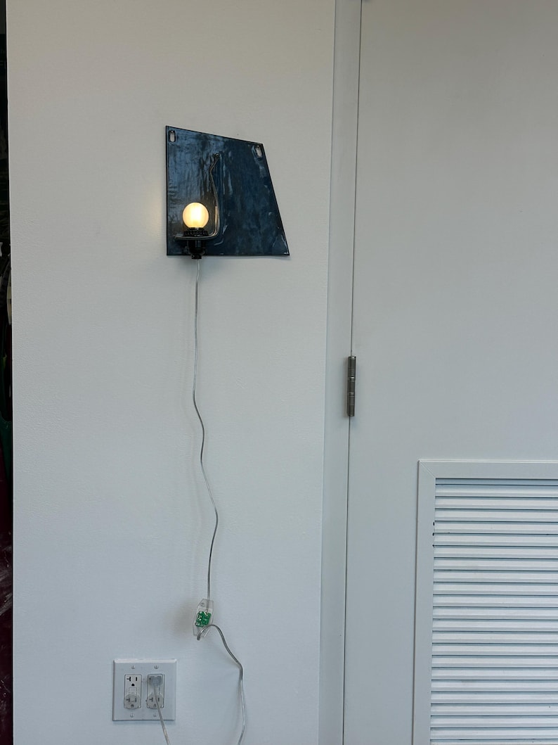 May include: A modern wall sconce with a dark blue, irregularly shaped metal backplate. A small, round light bulb is mounted on a black fixture. The sconce is connected to a wall outlet via a gray cord.