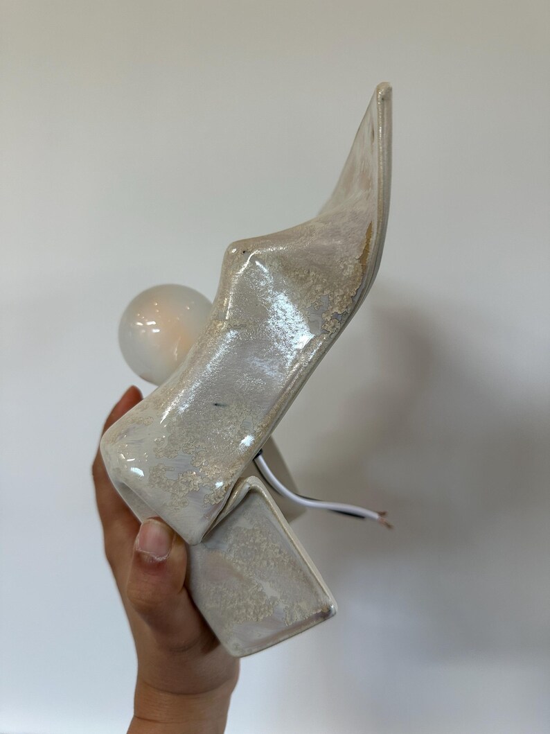 May include: A sculptural ceramic wall lamp with a glossy, mottled off-white and beige glaze. The fixture features an angular, pointed silhouette with a protruding circular white light bulb socket and exposed electrical wiring extending from the base.