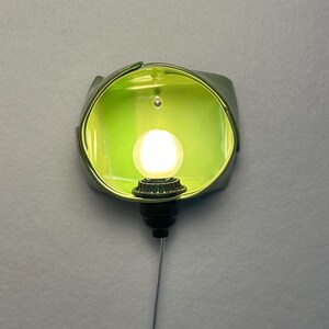 May include: A wall-mounted light fixture featuring a lime green, bowl-shaped ceramic shade with a glossy finish. A single, round, glowing white light bulb is centered within the shade, attached to a black socket with a thin, dangling power cord.