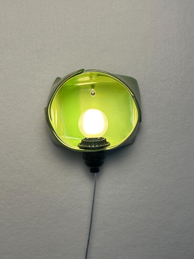 May include: A wall-mounted light fixture featuring a lime green, bowl-shaped ceramic shade with a glossy finish. A single, round, glowing white light bulb is centered within the shade, attached to a black socket with a thin, dangling power cord.