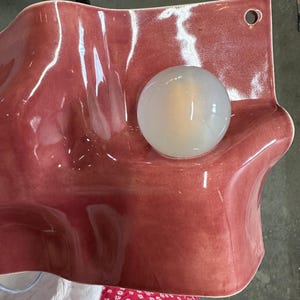 May include: A wall-mounted ceramic light fixture with a glossy, wavy, dusty rose-colored surface. The piece features a circular, translucent white glass globe bulb centered on the undulating form and a small hole in the top right corner for mounting.