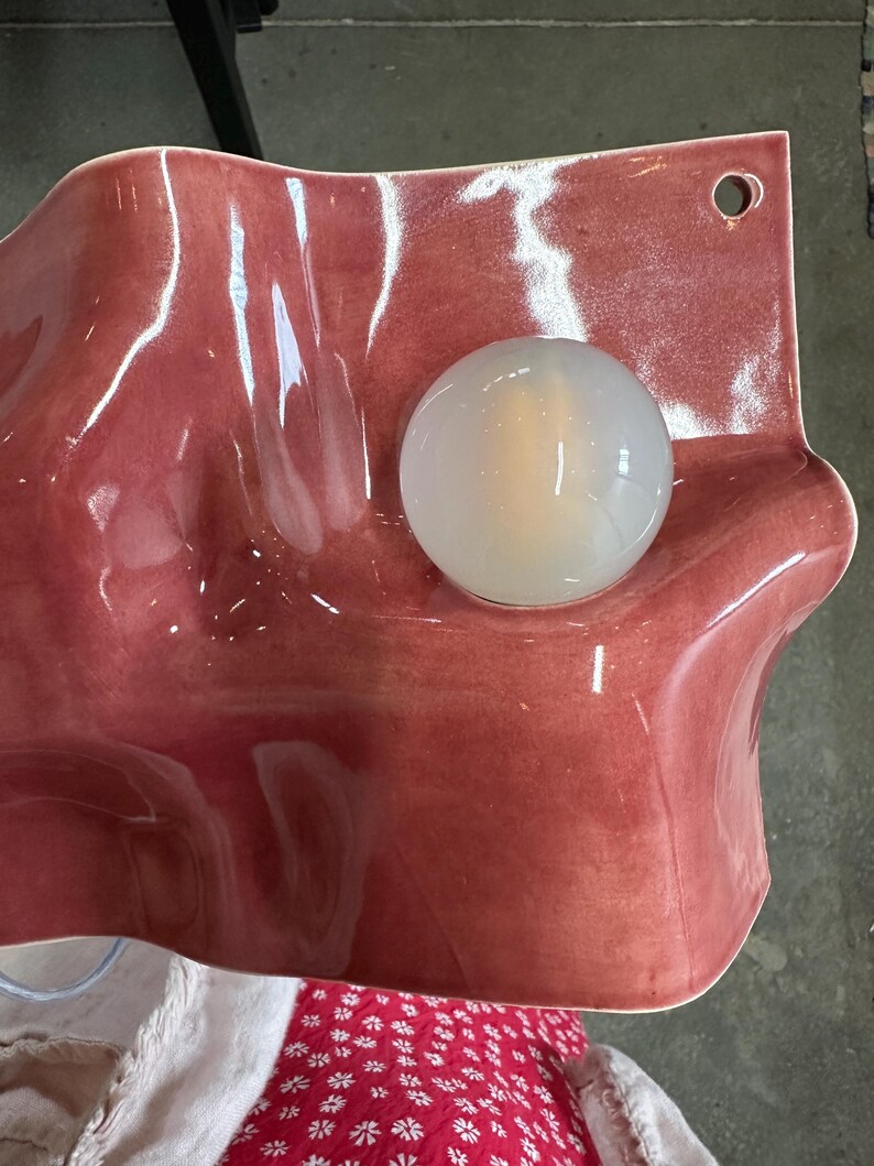 May include: A wall-mounted ceramic light fixture with a glossy, wavy, dusty rose-colored surface. The piece features a circular, translucent white glass globe bulb centered on the undulating form and a small hole in the top right corner for mounting.