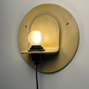 Pu&ograve; includere: Un'applique circolare in ceramica gialla dotata di un piccolo ripiano integrato che sostiene un portalampada nero e una lampadina sferica satinata accesa. L'apparecchio include due piccoli fori di montaggio laterali e un cavo di alimentazione nero.