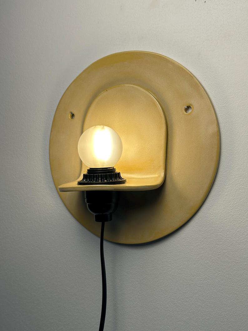 Pu&ograve; includere: Un'applique circolare in ceramica gialla dotata di un piccolo ripiano integrato che sostiene un portalampada nero e una lampadina sferica satinata accesa. L'apparecchio include due piccoli fori di montaggio laterali e un cavo di alimentazione nero.