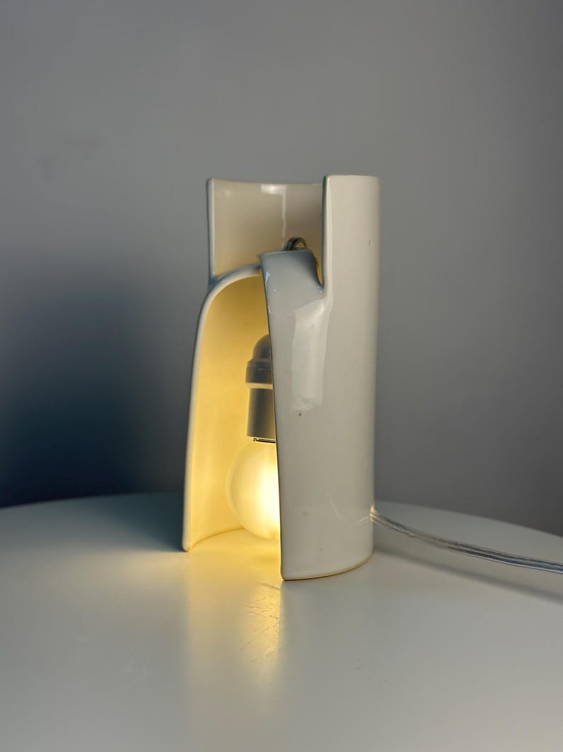 May include: A white ceramic table lamp with a curved, cylindrical structure that features an open archway. A single light bulb is positioned inside the hollow center, emitting a warm glow. A thin, clear power cord extends from the base of the lamp.