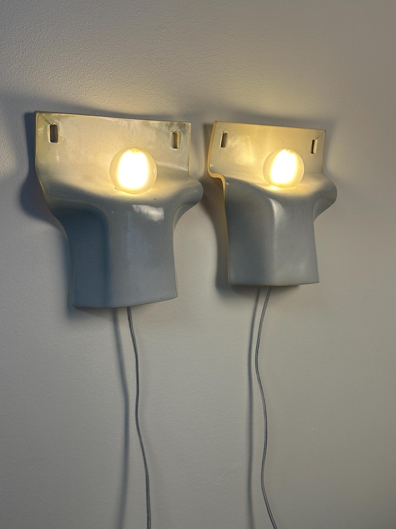 May include: A pair of wall-mounted light fixtures featuring a smooth, pale blue ceramic body with a curved, sculptural form. Each fixture holds a single glowing, spherical light bulb and includes two rectangular mounting holes at the top.