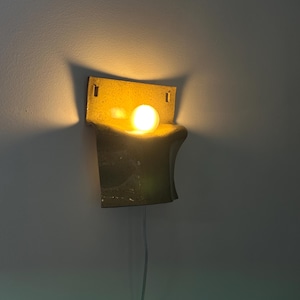 May include: A wall-mounted sconce featuring a curved, speckled yellow ceramic shade with a rectangular backplate. The fixture holds a single glowing, warm-toned spherical light bulb that casts a soft illumination against the wall surface.