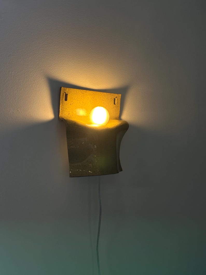 May include: A wall-mounted sconce featuring a curved, speckled yellow ceramic shade with a rectangular backplate. The fixture holds a single glowing, warm-toned spherical light bulb that casts a soft illumination against the wall surface.