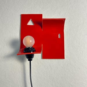 May include: A contemporary red wall light with a geometric design. It has a triangular cut-out and a small, round light bulb. The bulb is mounted on a black base, with a black cord hanging down. The light is designed for indoor use.