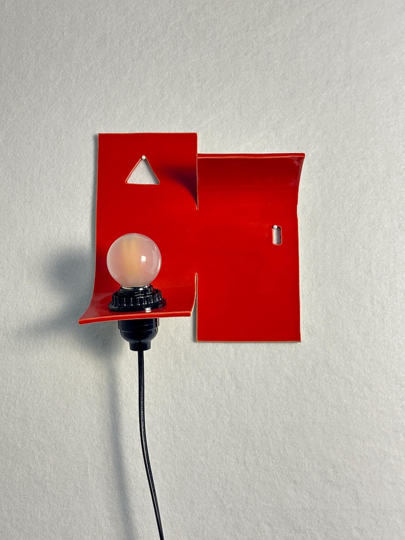 May include: A contemporary red wall light with a geometric design. It has a triangular cut-out and a small, round light bulb. The bulb is mounted on a black base, with a black cord hanging down. The light is designed for indoor use.