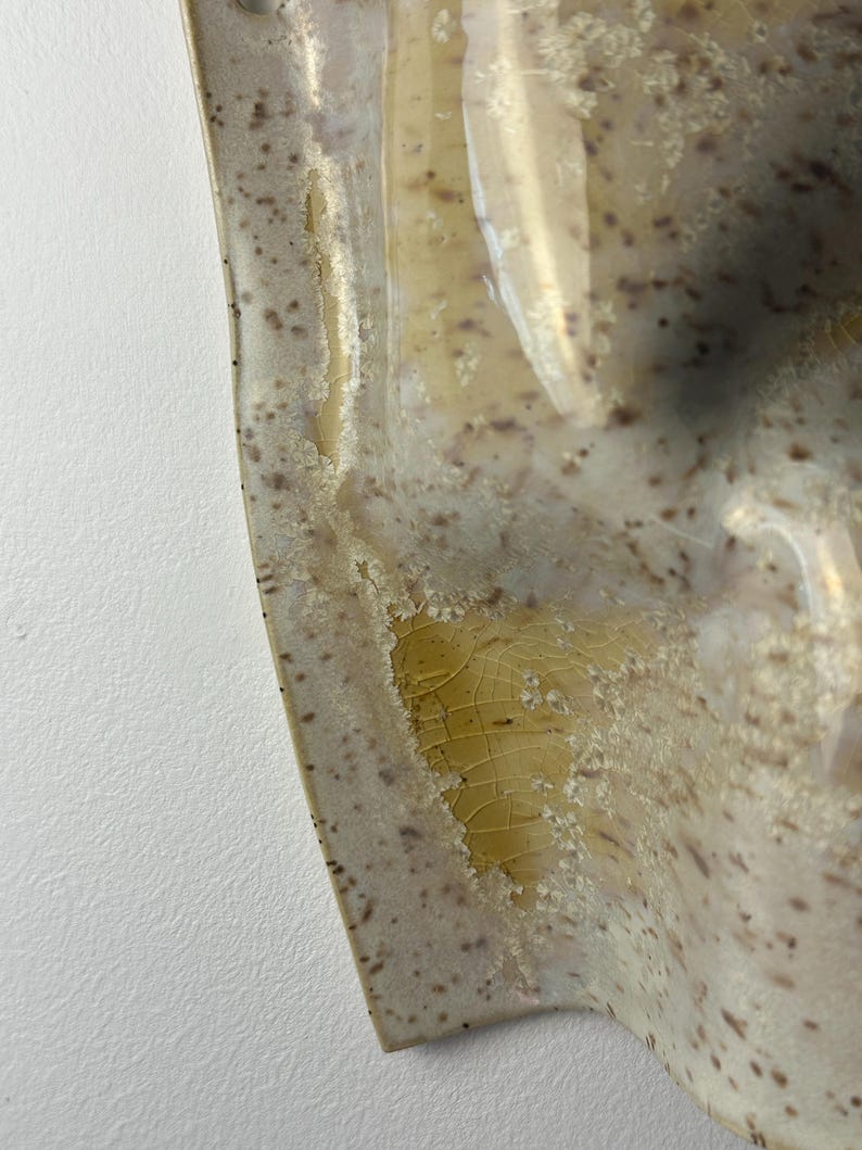 May include: A ceramic wall piece with a curved, organic shape featuring a speckled cream and beige surface. The glaze exhibits a distinct crackle texture with translucent amber-colored pooling in the recessed areas and dark brown speckles.