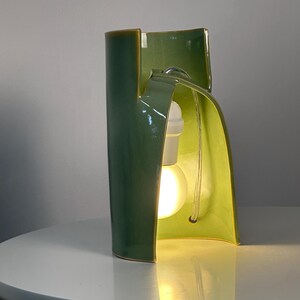 May include: A sculptural ceramic table lamp with a glossy sage green exterior and a lighter lime green interior. The curved, organic form features a central cutout housing a white light socket and a glowing round light bulb with a clear power cord.