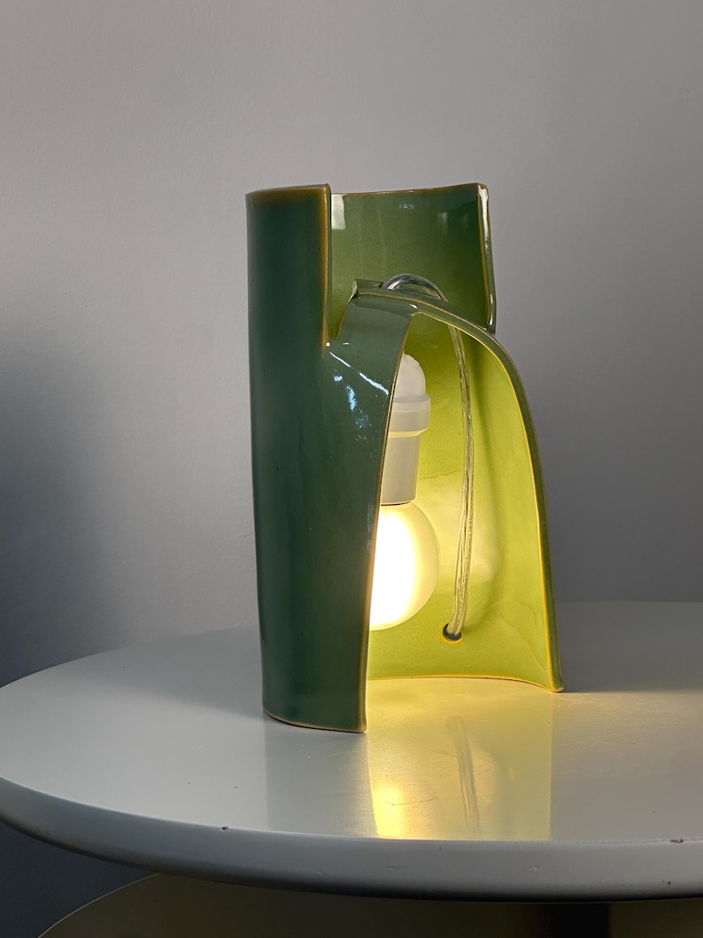 May include: A sculptural ceramic table lamp with a glossy sage green exterior and a lighter lime green interior. The curved, organic form features a central cutout housing a white light socket and a glowing round light bulb with a clear power cord.